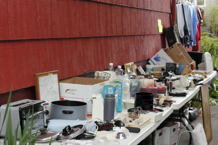 Variety of old, unwanted objects on two tables near a wall of a house in a driveway being displayed for sale at a yard sale.