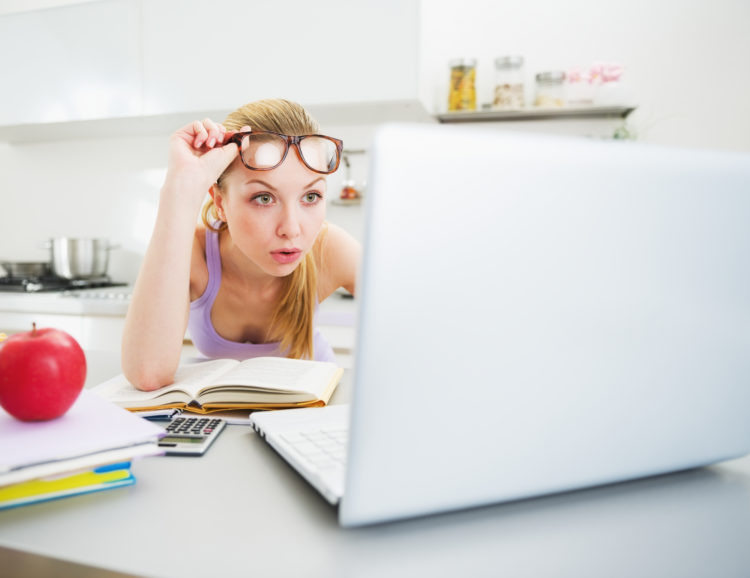 Image of surprised young woman looking in laptop while studying in kitchen