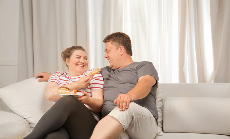 Image of Overweight couple eating sweets on sofa at home